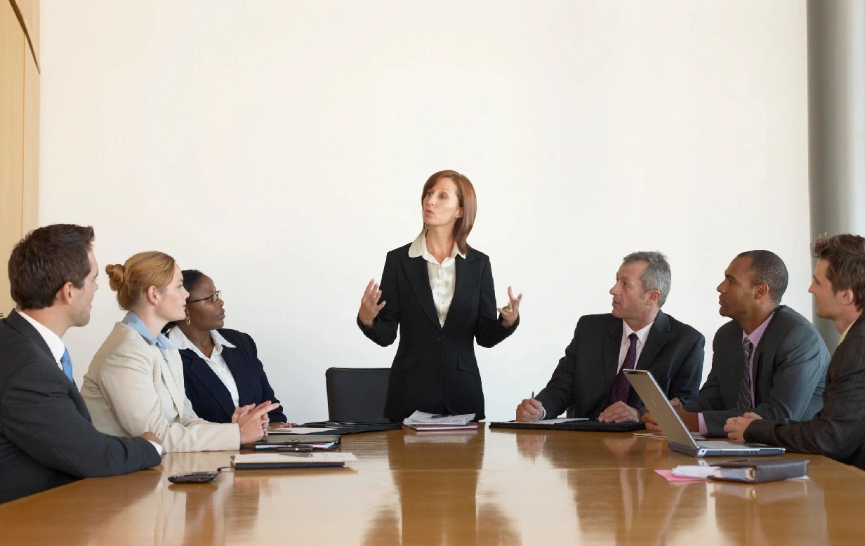 A woman standing at the head of a table with four other people.